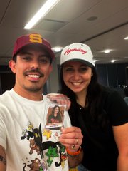 A smiling man wearing a brown cap and white graphic t-shirt stands next to a smiling woman in a white "Royals" cap and black shirt, who is holding up a small packaged keychain.