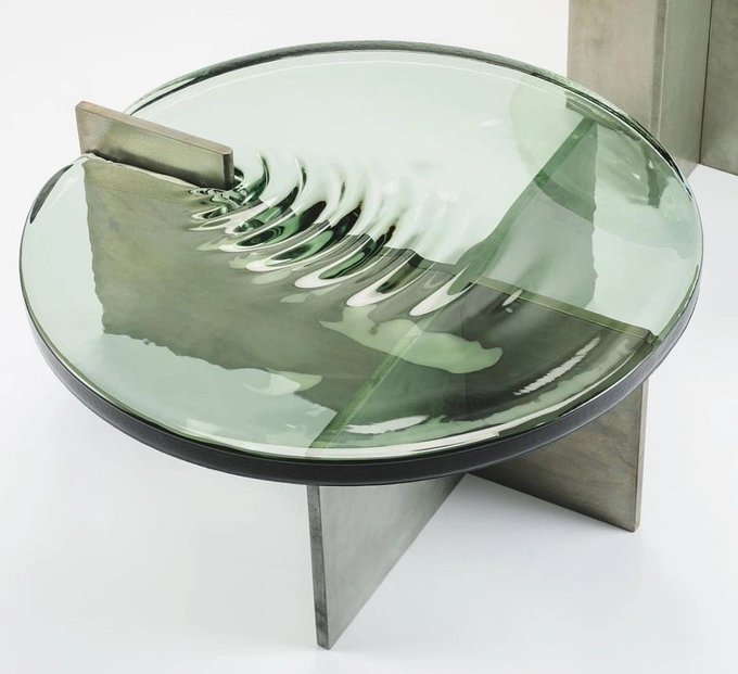 A sculptural art piece featuring a circular glass dish with ripple-like distortions, mounted on angular metal supports against a white background.