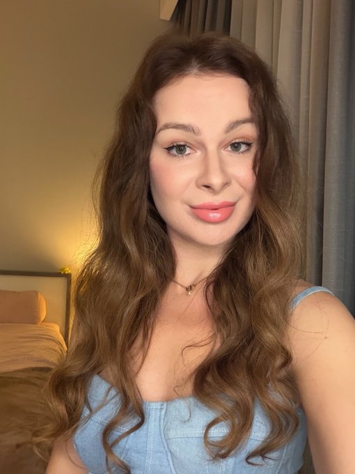 A selfie of a woman with long wavy brown hair wearing a light blue sleeveless top and a small heart necklace, smiling softly in a bedroom setting.