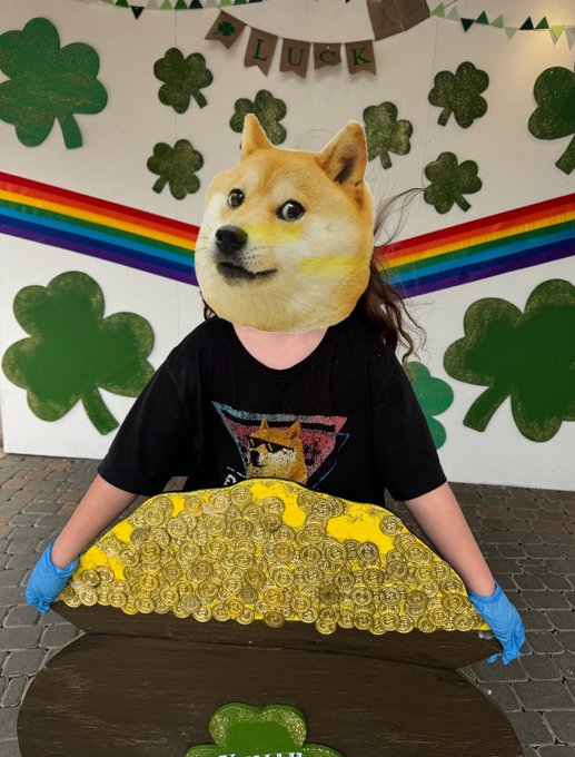 A person wearing a Shiba Inu (Doge) mask and black t-shirt, holding a pot of gold coins in front of a St. Patrick's Day-themed backdrop with shamrocks, a rainbow, and a "LUCK" banner.