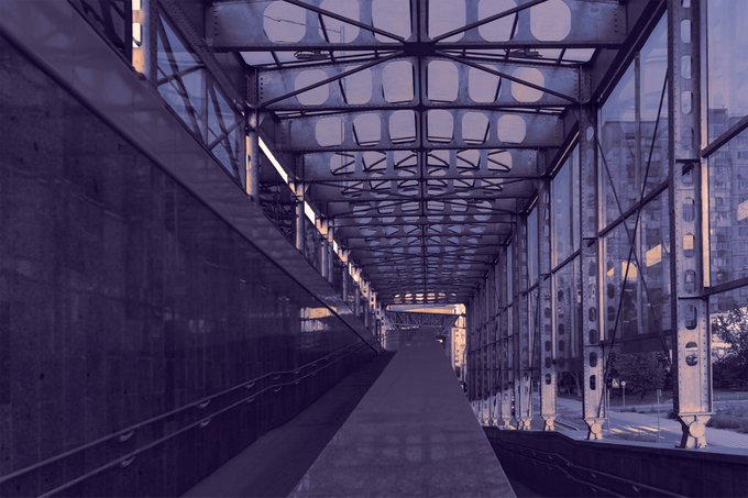 A view looking through an industrial steel and glass bridge or walkway with overhead trusswork, with a bluish tint over the entire scene.
