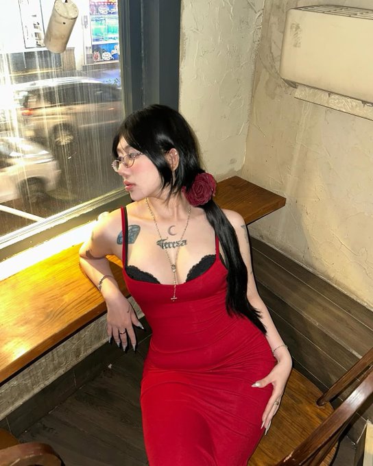 A woman with long black hair, glasses, and tattoos sits by a wooden bench in a red dress with a black lace bra, wearing a necklace and a red rose in her hair.