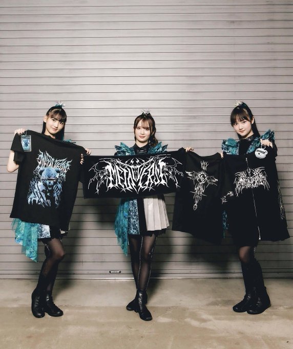 Three young women in matching teal and black gothic-style outfits holding up band merchandise (t-shirts and a banner) with stylized metal logos.