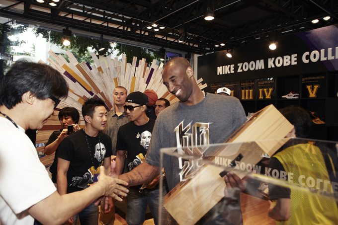 Kobe Bryant wearing a "24" shirt at a "NIKE ZOOM KOBE COLLECTION" event, shaking hands with a fan while holding a shoebox.