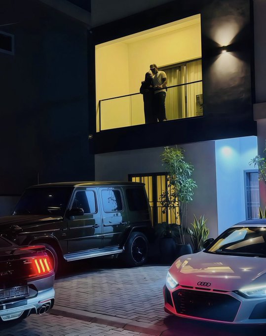 A nighttime scene showing a couple embracing on a lit balcony above a driveway where a black Mercedes G-Wagon and a white Audi are parked.