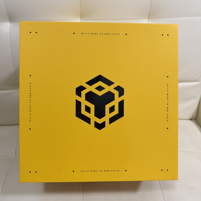 The image features a yellow box with black text, which appears to be a product or packaging for a company named @BNBCHAINZH. The box has a logo of a lion's head on it, indicating that it might be related to the company's branding or products. The box is placed on top of a white couch, suggesting that it could be a gift or promotional item from the company.