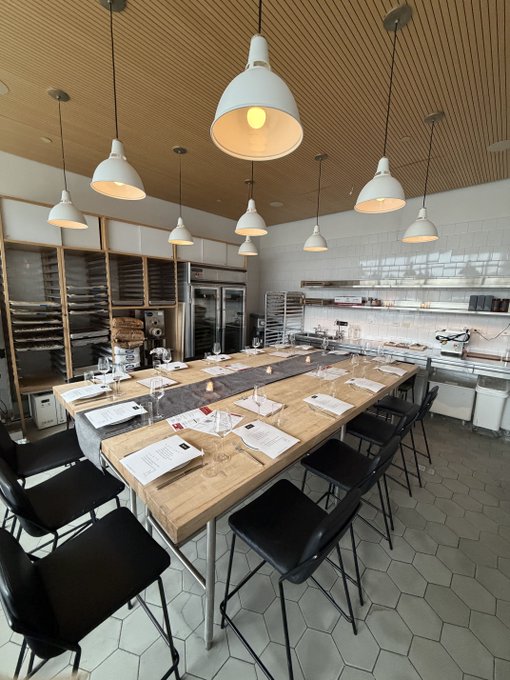 A modern, well-lit kitchen or culinary classroom features a long wooden table set with papers, surrounded by black chairs, under pendant lights and a wood-slatted ceiling with hexagonal floor tiles.