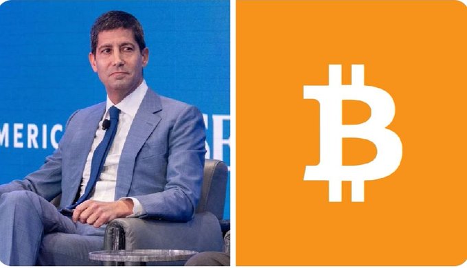 The image features a man sitting in a chair with his legs crossed, wearing a suit and tie. Behind him, there's a large orange square with the letter "B" written on it. The background of the image also includes text that reads "BIG: Fed Chair Kevin Warsh: Bitcoin is an important asset, and it doesn't trouble me." This suggests that the man in the suit might be involved in or interested in cryptocurrency, specifically Bitcoin.