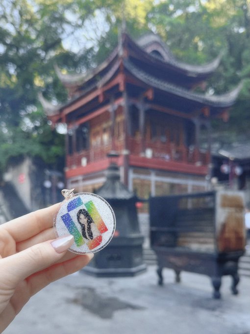 A hand holds up a round silver pendant featuring a colorful rainbow design and a figure, with a traditional Asian temple featuring curved roofs and incense burners visible in the background.