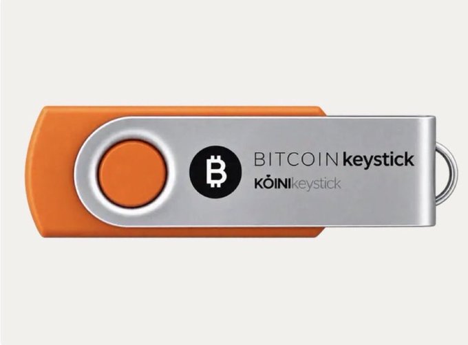 A product photograph of a metallic and orange 'BITCOIN keystick' USB drive made by KoinKeystick, displayed against a light background.