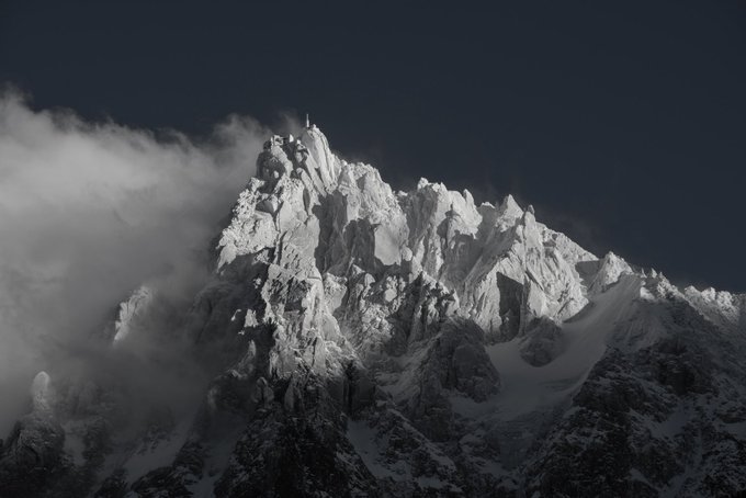A dramatic black and white photograph of a jagged, snow-covered mountain peak partially obscured by clouds against a dark sky.