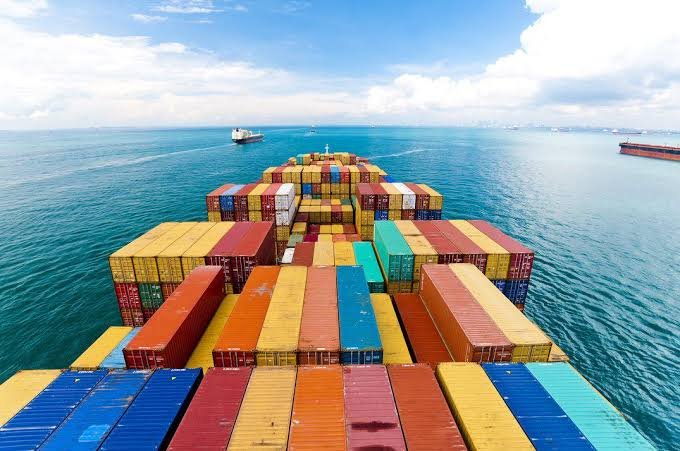 An aerial view from the bow of a container ship loaded with colorful cargo containers, with other vessels visible on the open ocean under a partly cloudy sky.