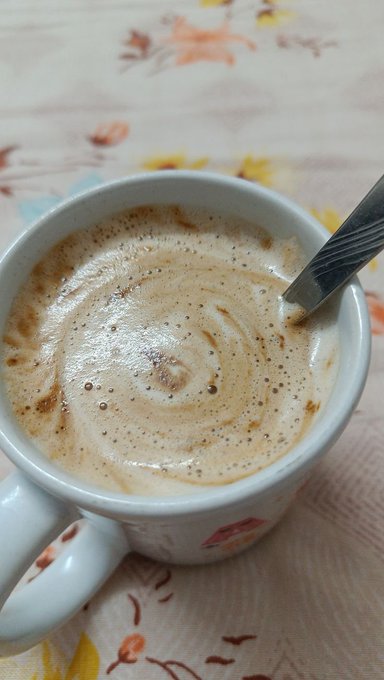 A close-up photograph of a white ceramic mug filled with a frothy brown coffee drink, resting on a floral patterned surface with a metal spoon inside.