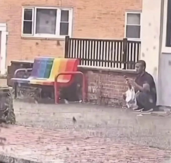 A person squats on a porch next to a colorful rainbow-striped plastic chair in front of a brick building.