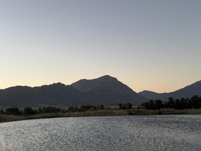 A scenic landscape showing a mountain range at dusk with its reflection on the calm water of a lake in the foreground.