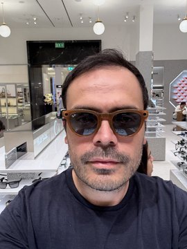 A selfie of a middle-aged man with short dark hair, stubble, and orange-tinted sunglasses, wearing a black t-shirt inside what appears to be a brightly lit retail store or showroom.