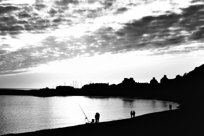 Black-and-white silhouette photograph of a beach scene at sunset, with people fishing on the shore and a town visible in the background across the water.