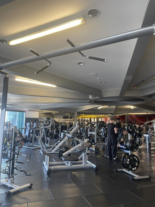 A gym interior with various weight machines, squat racks, and a pull-up bar overhead, with two people in the background.