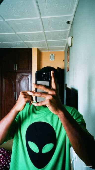 A person wearing a green alien t-shirt takes a mirror selfie with their phone, capturing a casual indoor setting with decorative ceiling tiles and wooden cabinets. The image's playful, laid-back vibe complements the post's message of patience and excitement for the @ProjectShiesty project.