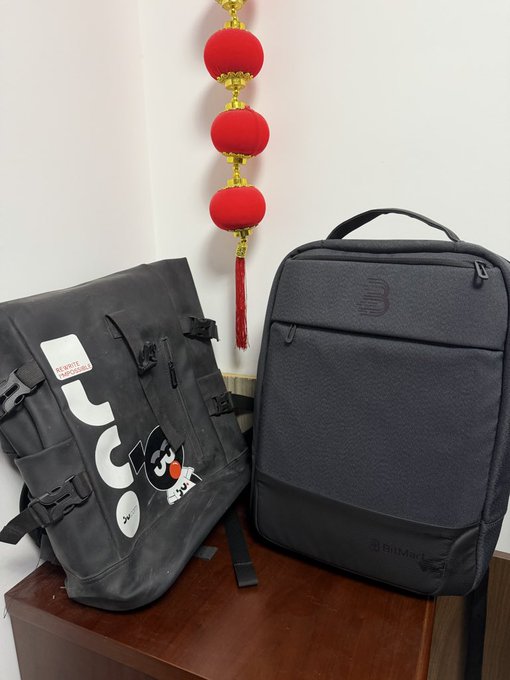 A black backpack with a 'UC' logo and various patches sits next to a dark grey Targus laptop bag on a wooden surface, with a red Chinese decorative lantern hanging in the background.