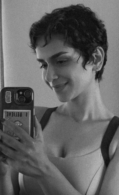 A black-and-white portrait of a person with short curly hair smiling while looking at a smartphone with a phone case displaying 'I BELIEVE' in mirrored text.