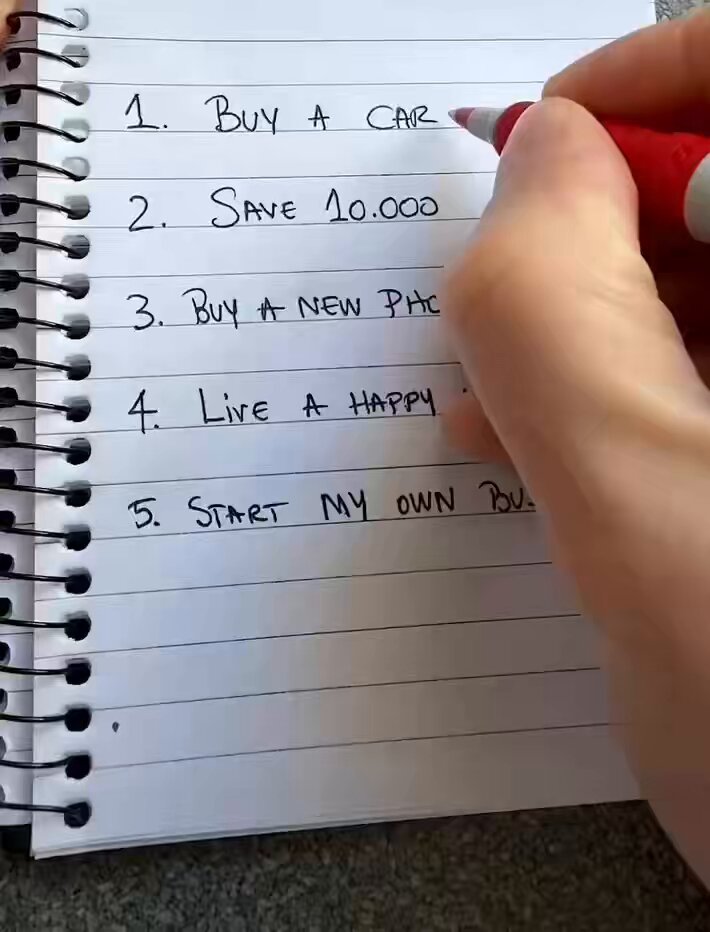 The image shows a hand writing on a piece of paper, which appears to be a to-do list or a shopping list for buying a car. The person's hand is holding a red pen as they write down their thoughts or tasks related to purchasing a new vehicle.