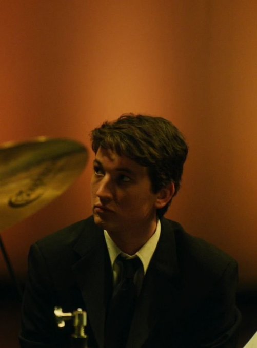 A moody, low-lit portrait of a young man in a dark suit looking off to the side, with a cymbal visible in the foreground, suggesting a jazz or music setting.