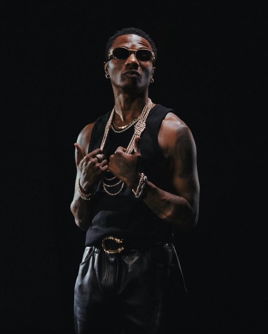 A man wearing sunglasses, a black tank top, and gold chains stands against a dark background, pointing to his chest with both hands.