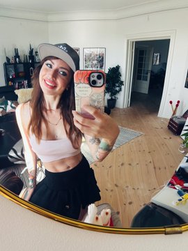 A fisheye mirror selfie of a smiling young woman wearing a white cap, pink tube top, and black skirt, taking a mirror selfie with a pink phone in a living room.
