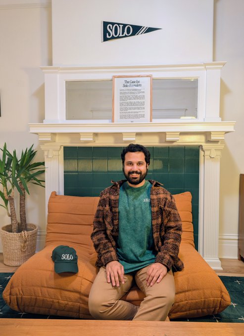 A smiling man with a beard sits on a large orange cushion in front of a fireplace, wearing a green 'SOLO' t-shirt and a patterned jacket, with a 'SOLO' pennant mounted above.