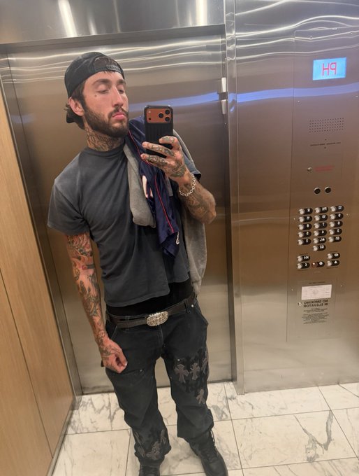 A tattooed man wearing a backwards cap, gray t-shirt, and dark jeans poses for a mirror selfie inside an elevator displaying "149." The image complements a Twitch livestream announcement promising to give away $25,000 in crypto to viewers who share their Solana addresses, fitting the casual, flashy aesthetic of a streaming giveaway promotion.