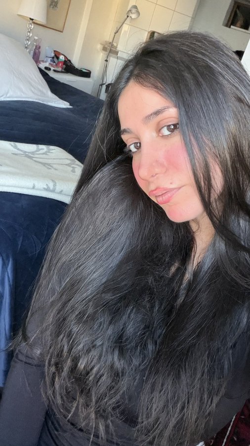 A close-up selfie of a young woman with long dark hair and flushed cheeks, looking at the camera in a bedroom setting with a bed visible behind her.