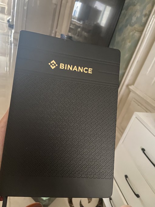 A person holds up a black box with the "BINANCE" logo and name printed in gold lettering.