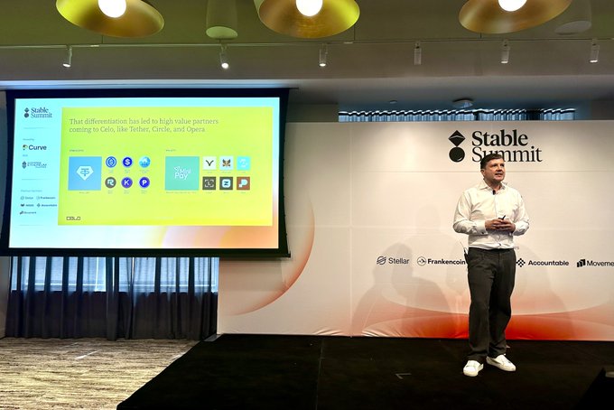 In this image, a man wearing a white shirt and gray pants stands on stage at an event, with a large screen behind him displaying information related to stablecoins. The screen shows various icons representing different aspects of the stablecoin ecosystem, such as logos for Bitcoin Core, Celo, and RippleXR. Additionally, there are other people in the image, likely attendees or participants of the event, positioned around the stage area. The context suggests that this might be a seminar or conference about stablecoins, with speakers presenting information on the potential benefits and applications of using stablecoins for daily currency transactions.