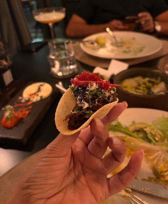 A hand holds a small taco topped with meat, greens, shredded cheese, and red garnish at a restaurant table with other dishes and a cocktail in the background.