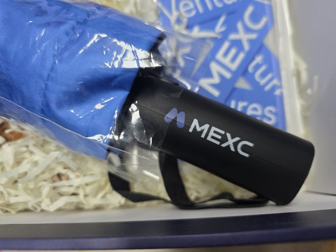 A black cylindrical case bearing the "MEXC" logo rests inside a gift basket filled with shredded paper and blue fabric, matching the MEXC-branded umbrella advertised in the background banner. The case likely contains the high-quality umbrella referenced in the post, which the recipient praised over the inferior umbrella that came with their Cullinan—prompting them to return the luxury car.