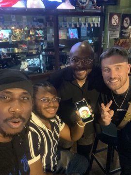 Four people pose together for a selfie at what appears to be an arcade or bar, with one person flashing a peace sign.