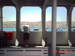 A scenic view from inside a ferry or boat, where a person stands looking out the windows at a coastal city and bay under a clear sky, with a date stamp of "08 08 2025" in the corner.