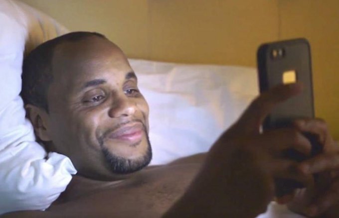A man lies in bed against white pillows, smiling as he looks at his smartphone.