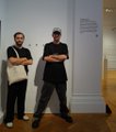 Two people stand posing in a gallery setting in front of a wall plaque.