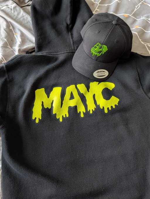 A black hoodie laid out on a bed, featuring the word "MAX" in bright yellow dripping letters, accompanied by a matching black "DEADBACK" snapback cap with a green skull logo.
