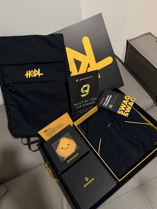 A Binance VIP swag box containing a black backpack with the BNB logo, a Binance VIP card, a plush yellow Binance mascot, black apparel, and a 'Built by You VIP Dinner' invitation card.