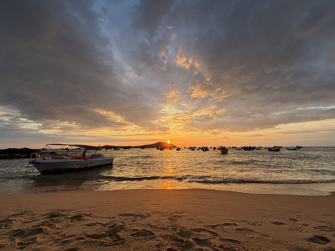 A sunset scene over a beach with boats anchored in calm water, golden sunlight reflecting off the ocean and scattered clouds in the sky.