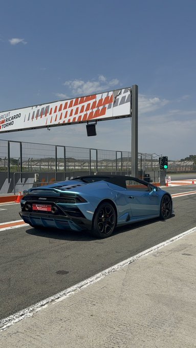 The image features a blue Lamborghini Huracan EVO sports car driving down an empty street with a track sign behind it, indicating that it's at a race track. The car appears to be in motion, possibly racing or participating in a practice session. In the background, there is a traffic light and a fence, suggesting that this location might be near a road intersection or a designated area for racing events.