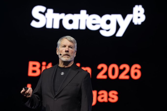 Michael Saylor stands on stage in a black suit with the 'Strategy' logo and 'Bitcoin 2026' text displayed on the screen behind him.