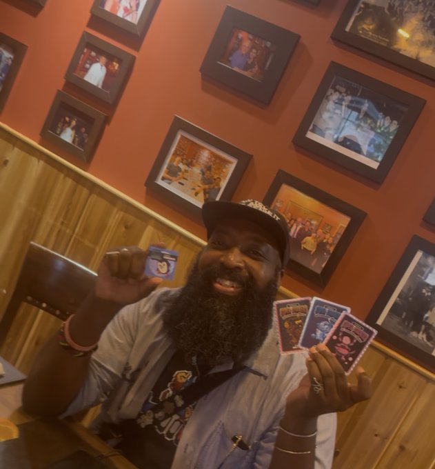A smiling bearded man wearing a cap poses happily in a restaurant with orange walls covered in framed photos, holding up tarot-style cards and a small card.