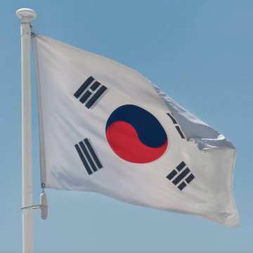 The South Korean national flag, known as the Taegeukgi, waves against a clear blue sky, displaying its white background, central red and blue Taegeuk symbol, and black trigrams in the corners.