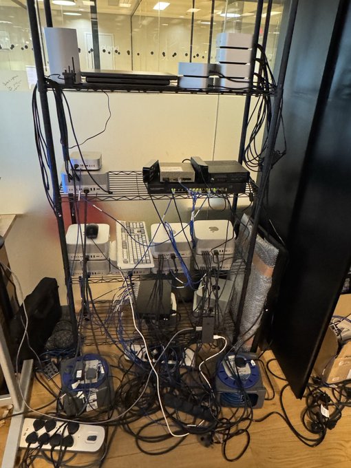 A tangled mess of cables, networking gear, and two routers sits on a metal wire shelving unit in what appears to be a lab or office setup, with the caption "Exo is cooking" humorously implying that the chaotic wiring job is a recipe in progress.