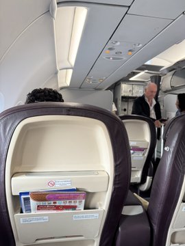 Interior view of an airplane cabin showing business class seats in burgundy with white headrests, with a flight attendant visible in the aisle.