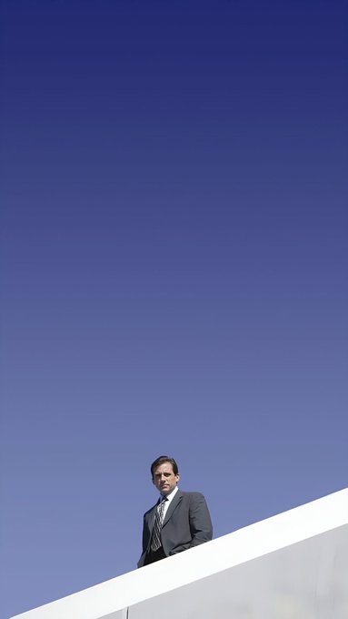 A low-angle photograph of a man in a dark suit standing at the top of a stark white structure against a clear blue sky.